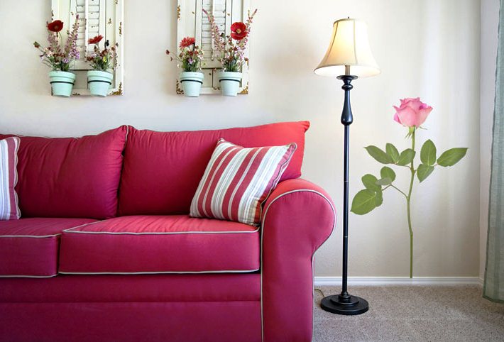 Красивый лиловый диванчик, роза на стене и красные цветы в горшочках идеально дополняют дизайн гостиной комнаты